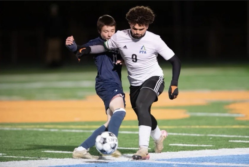 Pine Tree Academy boys varsity soccer player number 9 in white challenging for the ball against a navy-jersey opponent under the lights